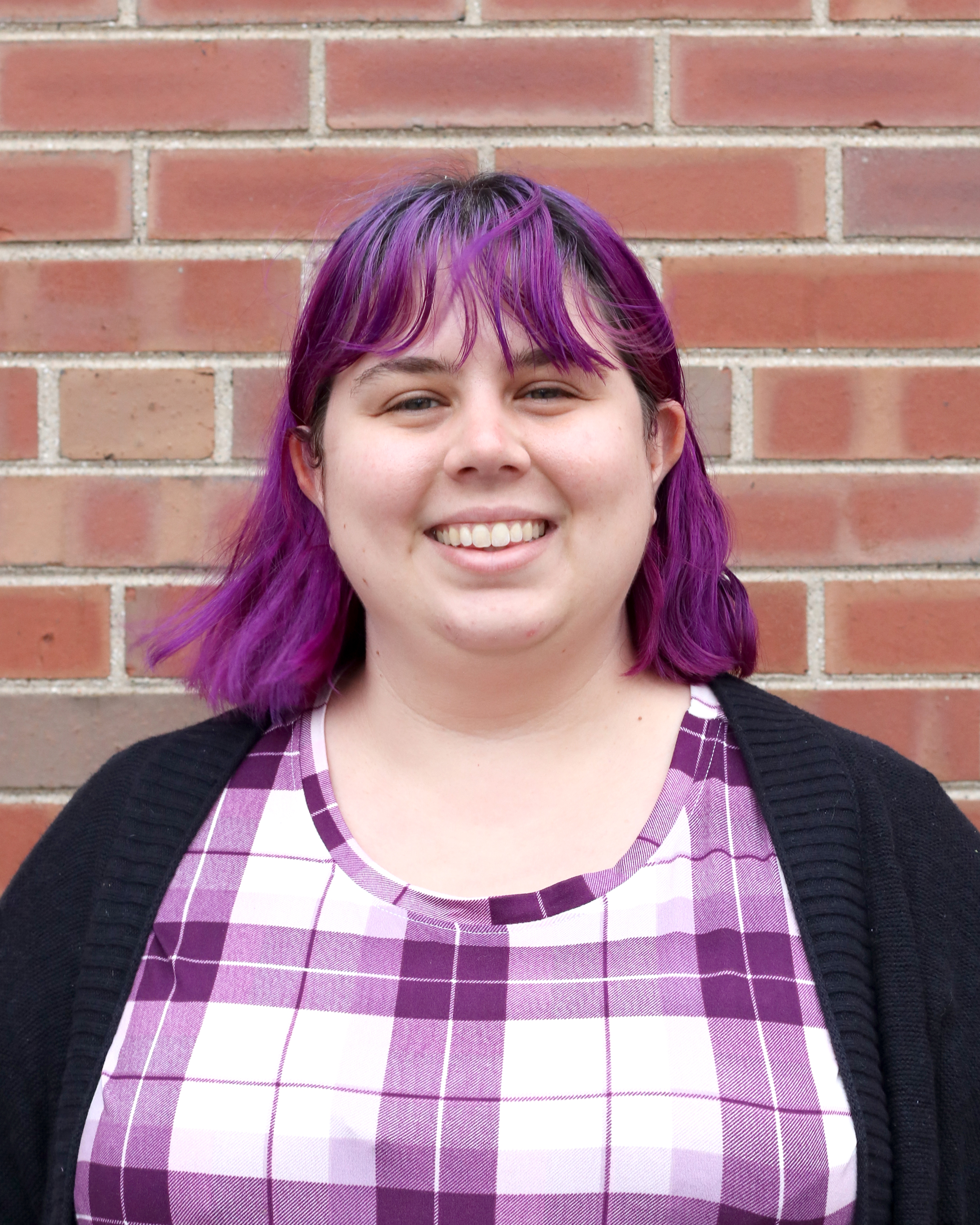 Jessie Bond, a white woman with purple hair and a purple plaid shirt, against a brick background
