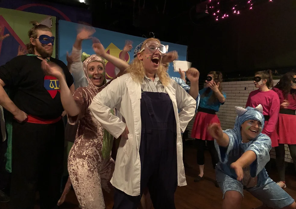 Cinderella, in a lab coat and safety goggles, smiles with her hands on her hips, the rest of the cast surrounding her