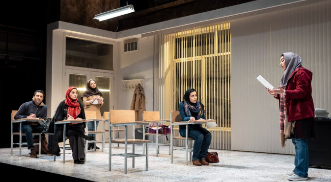 A woman in a hijab reads from a piece of paper to a classroom of other adults