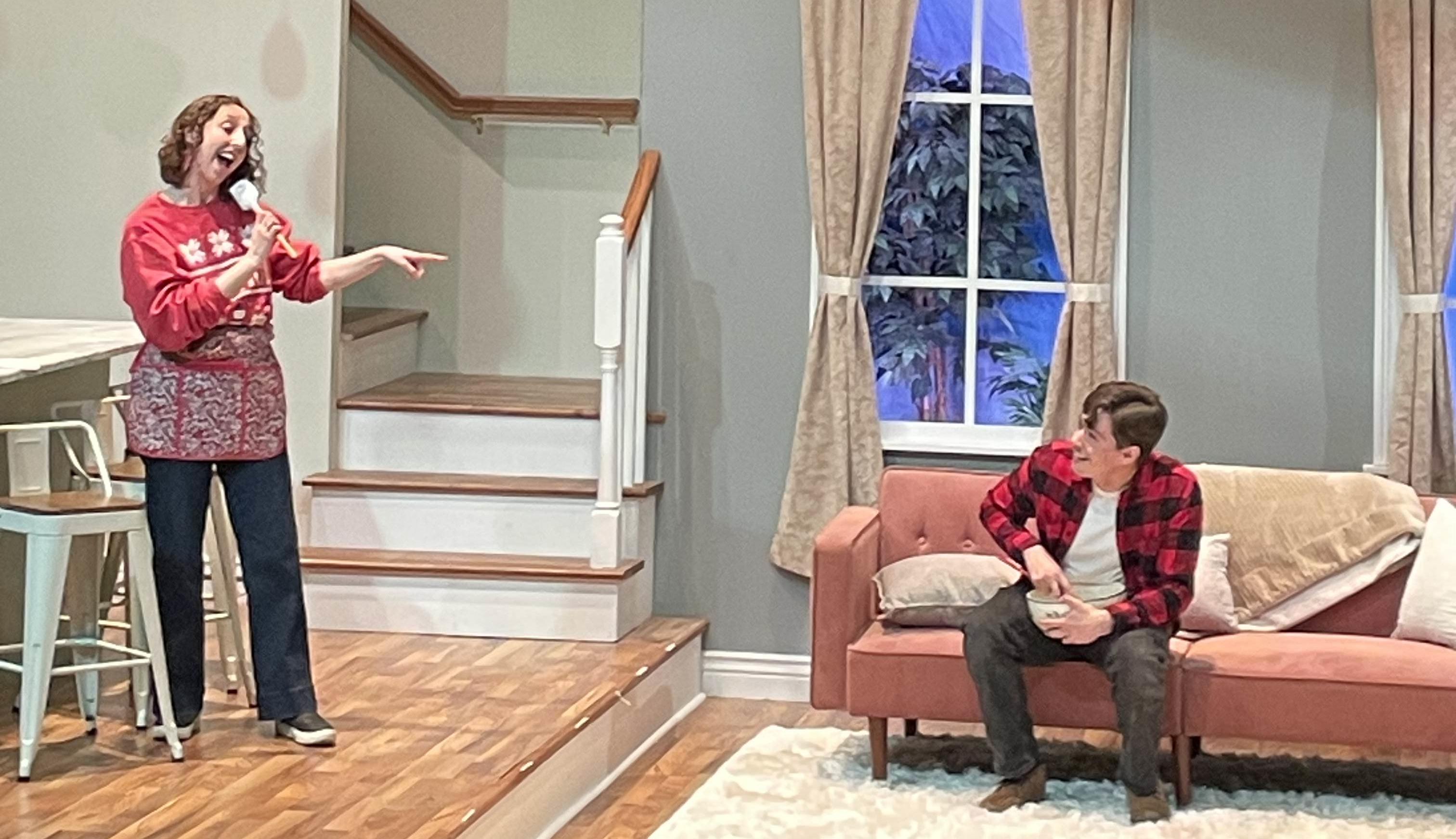 A woman in a red Christmas sweater and apron sings into a baking spatula and points at a smiling man sitting on a couch