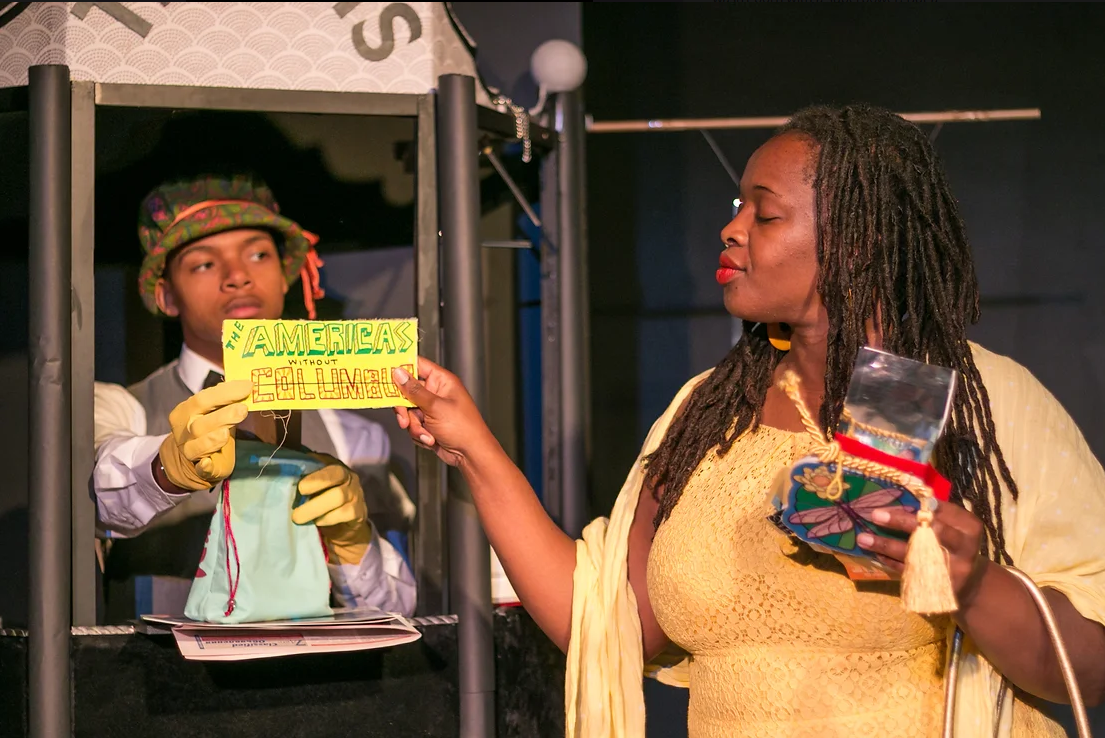 A man in a ticket booth hands a woman a ticket that reads The Americas without Columbus